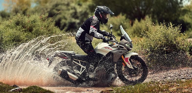 A TVS Apache RTX motorcycle in white color being ridden through a shallow stream, splashing water.